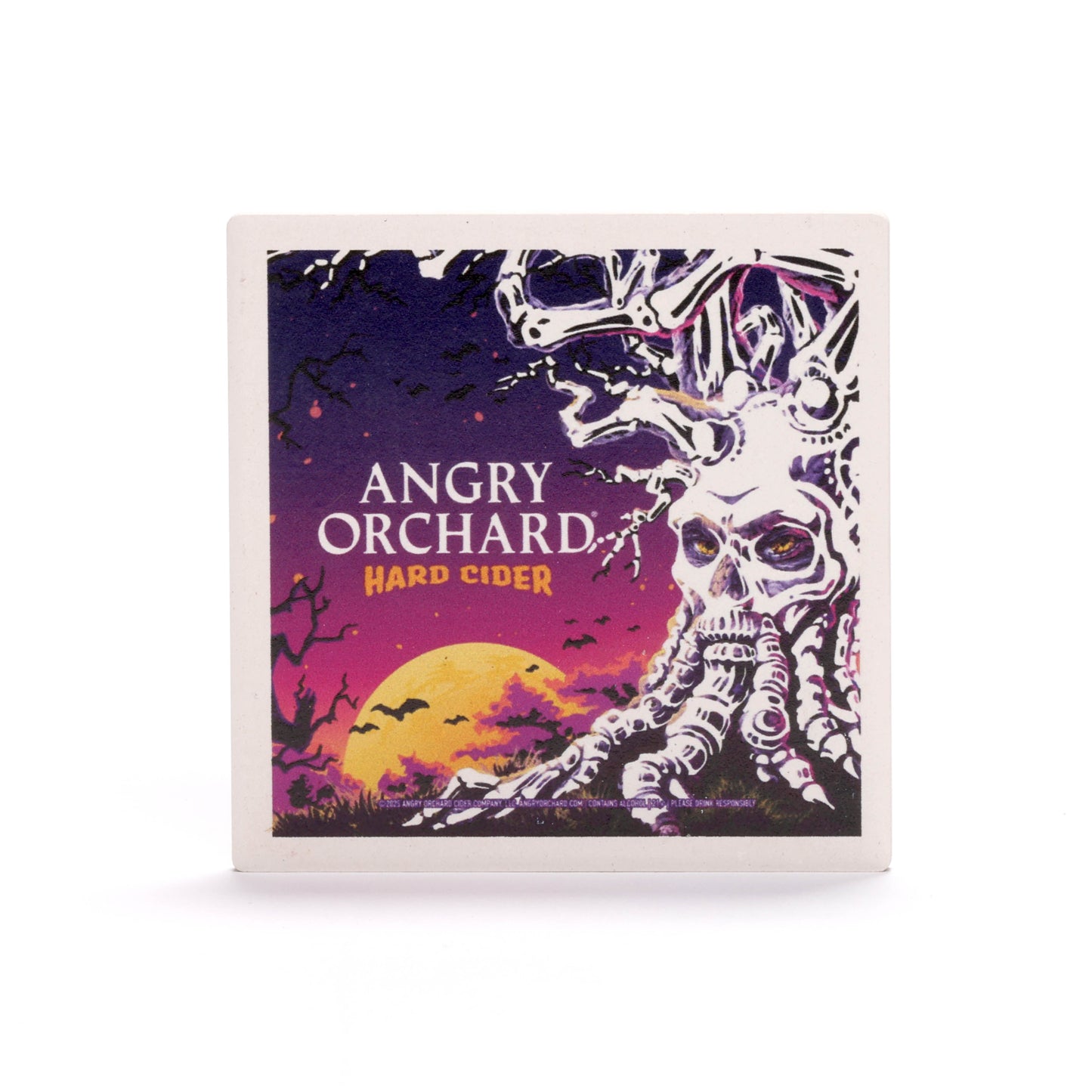 A square coaster with a Halloween-themed design. In the foreground, a gnarled tree with a skull-like face and boney branches stands against a purple and orange sky. A full yellow moon is visible in the background, with black bats flying across it. The text "ANGRY ORCHARD HARD CIDER" is written in white and orange letters to the left of the tree.